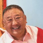 Een foto van een schuin naar links hellende Gelek rimpoche met een mooie lach met een rose blouse aan met een rode deken over zijn linker schouder. Op de achtergrond een blauwe muur.