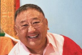 Een foto van een schuin naar links hellende Gelek rimpoche met een mooie lach met een rose blouse aan met een rode deken over zijn linker schouder. Op de achtergrond een blauwe muur.