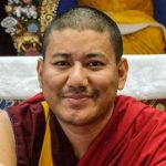 Een foto van een minzaam kijkende Demo Rinpoche met op de achtergrond het altaar van Jewel Heart Ann Arbor.