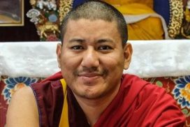 Een foto van een minzaam kijkende Demo Rinpoche met op de achtergrond het altaar van Jewel Heart Ann Arbor.