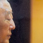 Een foto met een zwarte achtergrond met aan de rechterkant een vertikale gele balk. Op de voorgrond de helft van het gezicht van Gelek Rimpoche van de zijkant af gezien die in gedachten verzonken lijkt.