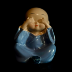 Je ziet een zwarte achtergrond met op de voorgrond een klein beeldje van een huilende boeddha ( met zijn handjes voor de ogen) met een blauw kleed aan.