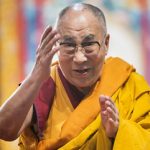 Een foto van de dalai lama die vol passie iets aan het vertellen is met zijn handen in gebarende houding.