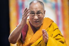Een foto van de dalai lama die vol passie iets aan het vertellen is met zijn handen in gebarende houding.